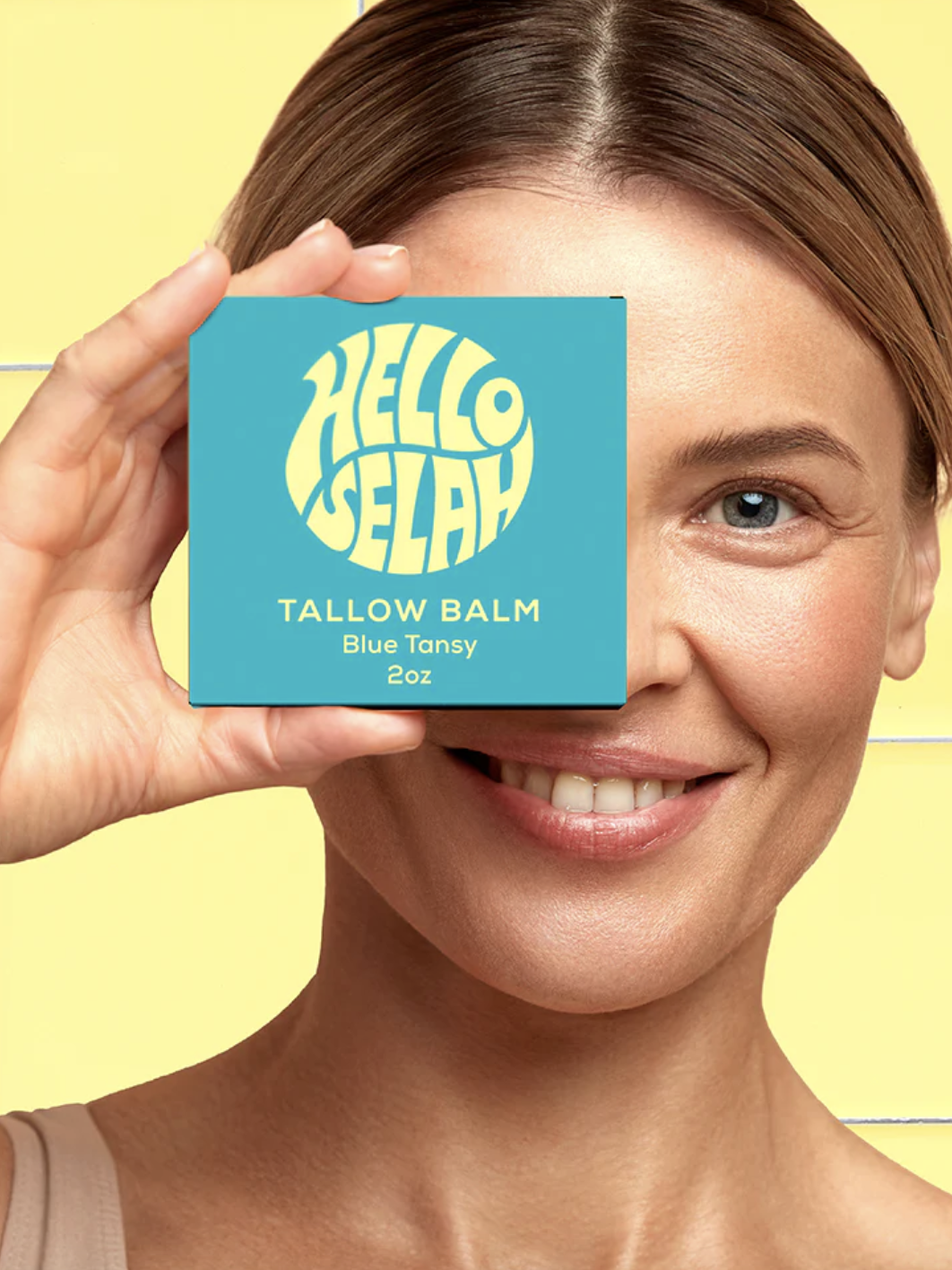 Smiling woman holding a "Hello Selah Tallow Balm" box over her eye against a yellow tiled background.