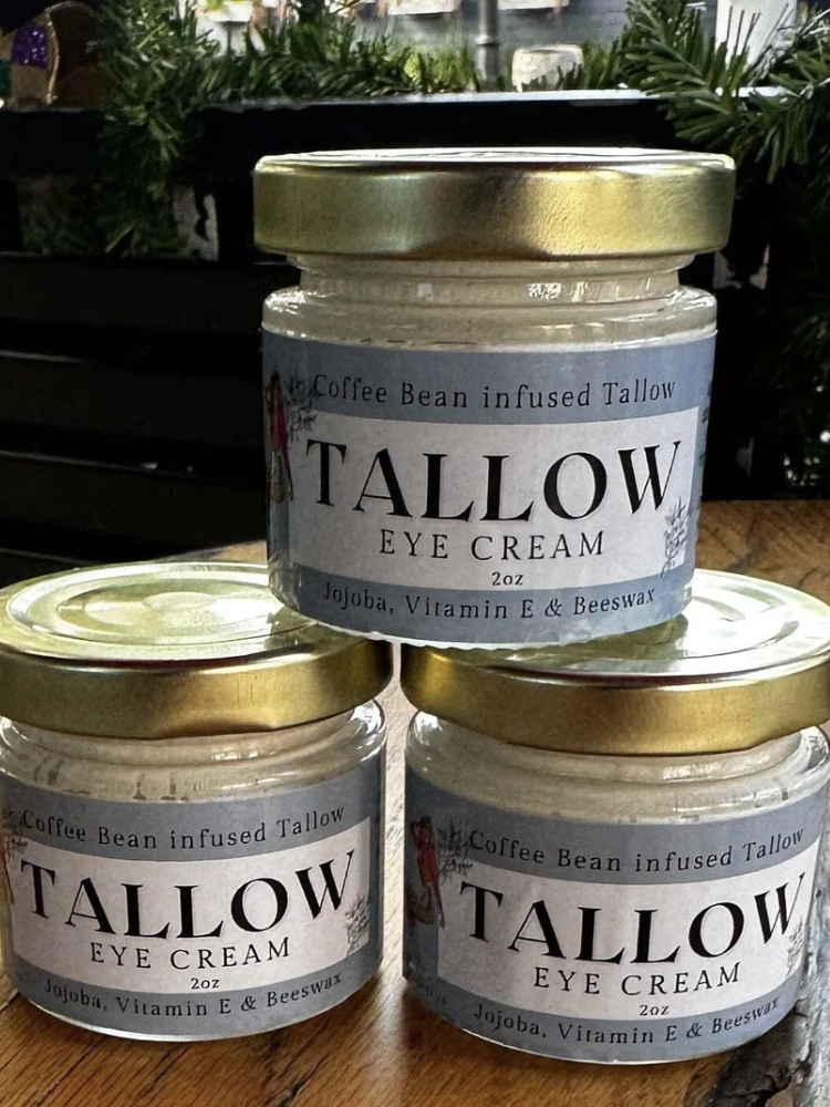 Three jars of Tallow Eye Cream, infused with coffee bean, jojoba, vitamin E, and beeswax, are stacked on a wooden surface with greenery in the background.