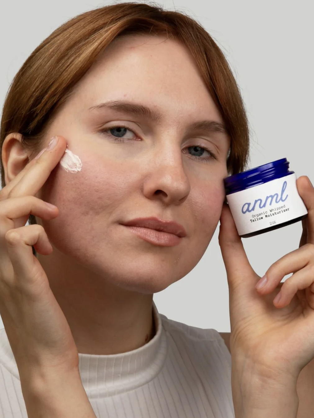 Person applying a cream labeled "anml" to their cheek, holding the container in their other hand.