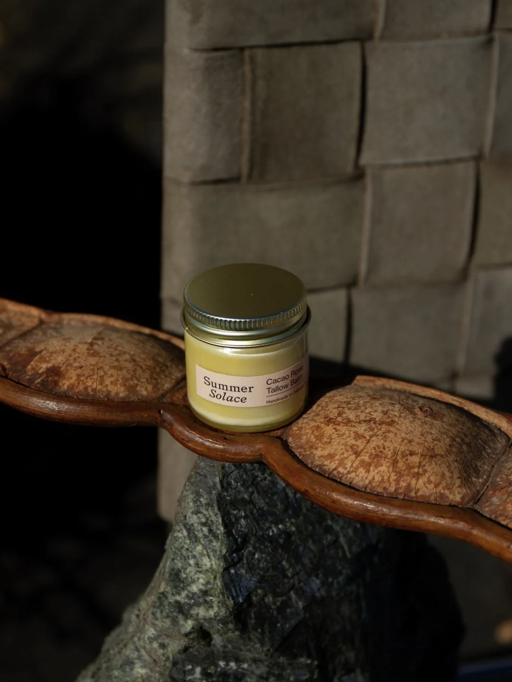 A jar labeled "Summer Solace" with cacao rose tallow, placed on a large seed pod, near a woven wall.
