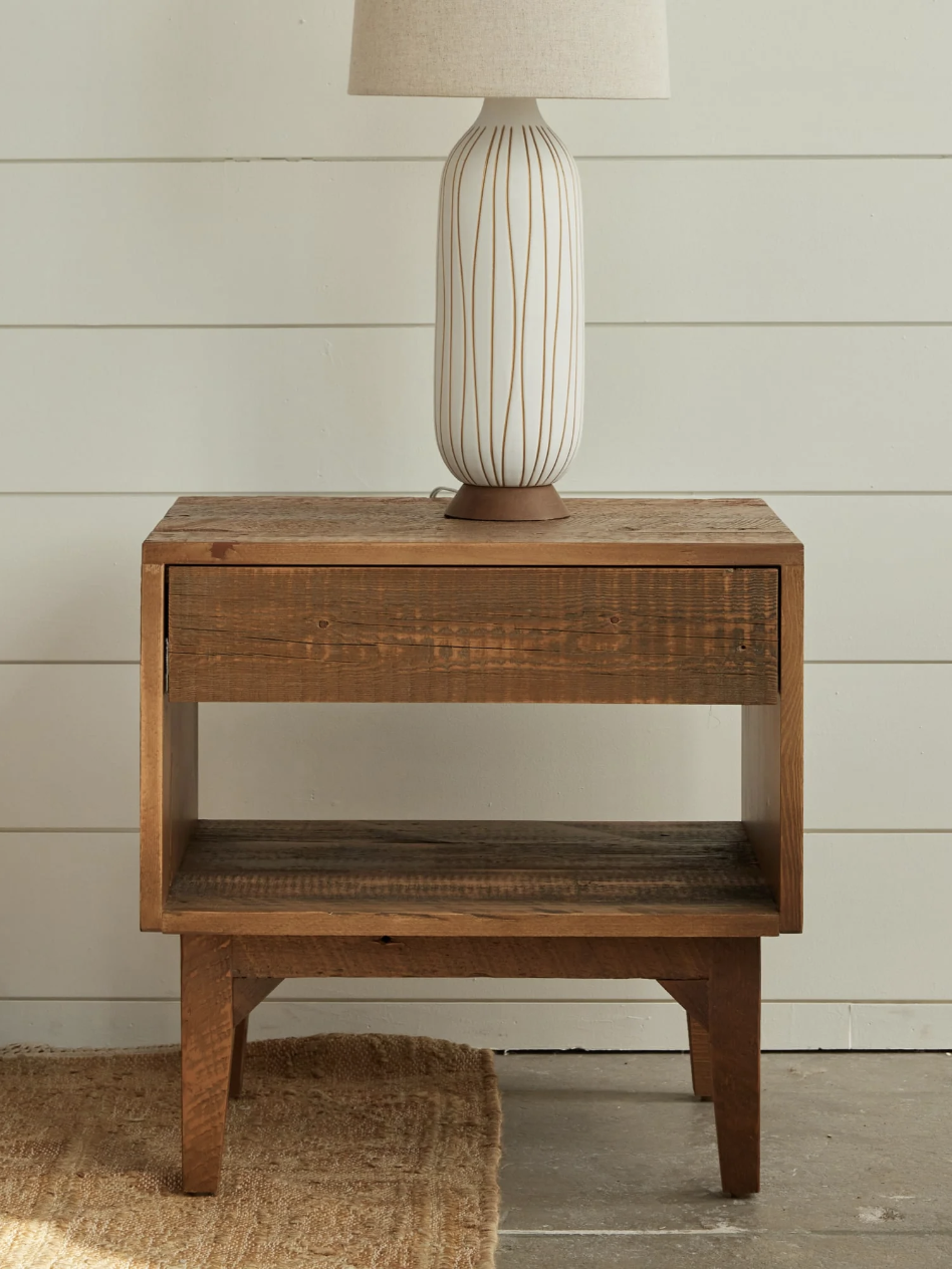 A wooden nightstand with a drawer and lower shelf, topped with a vertical-striped white lamp. The setting includes a beige rug on a concrete floor and a white panel wall background.