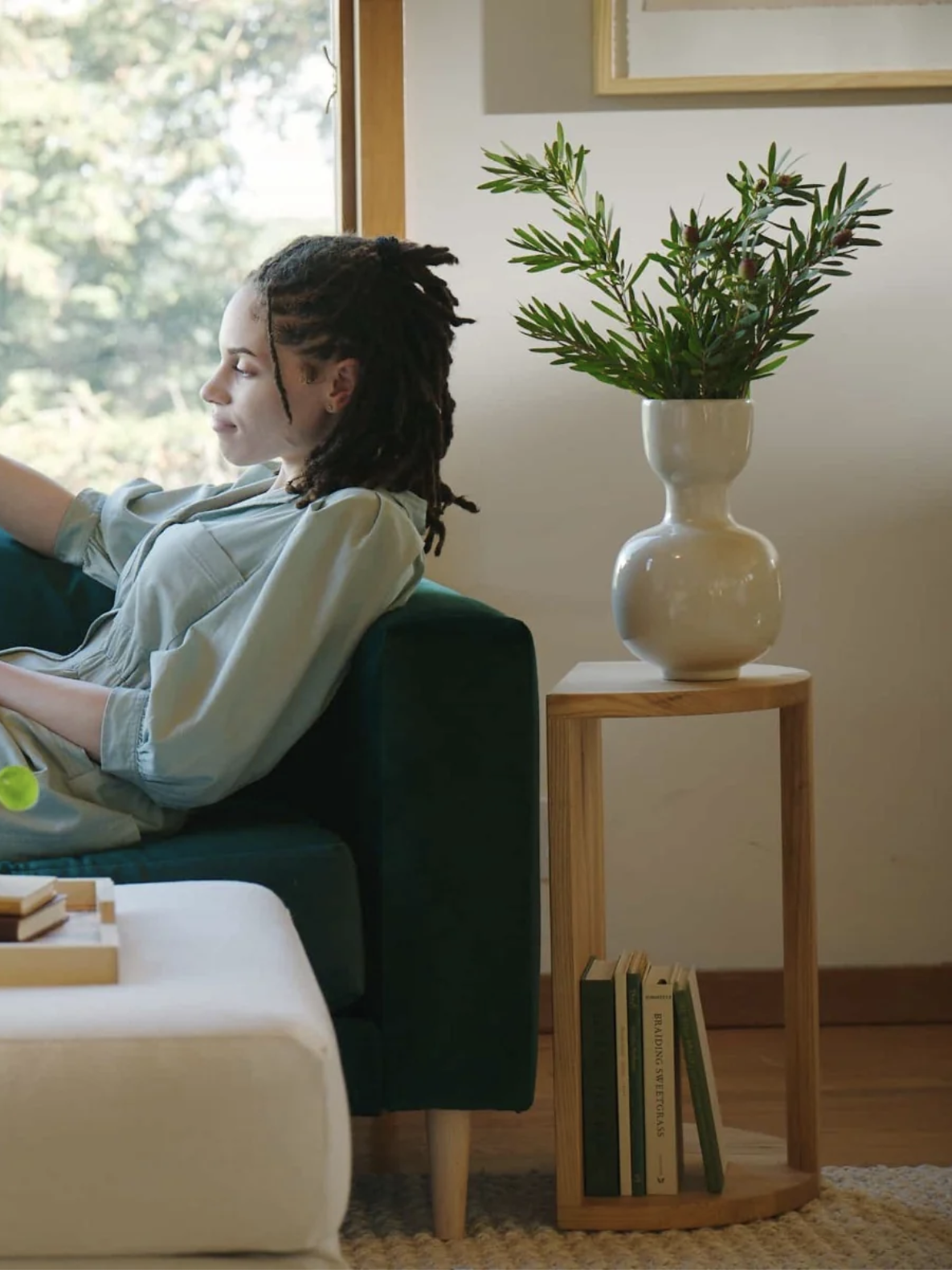 Person sitting on a dark green sofa near a window, next to a wooden table holding a white vase with green foliage and a few books underneath.