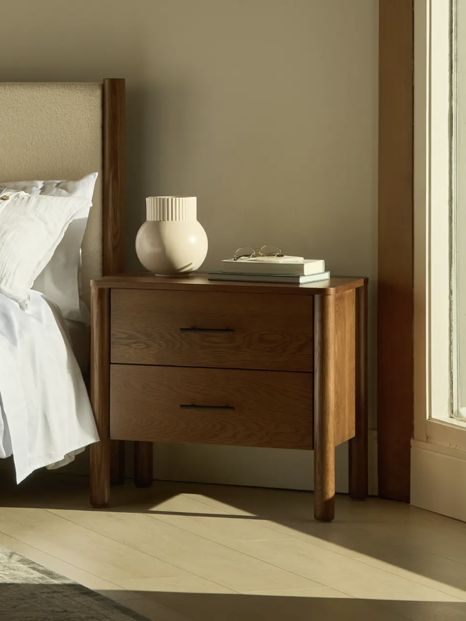 Wooden bedside table with drawers next to a bed with white sheets and a light brown headboard; a vase and books are on the table. Sunlight is streaming through a large window.