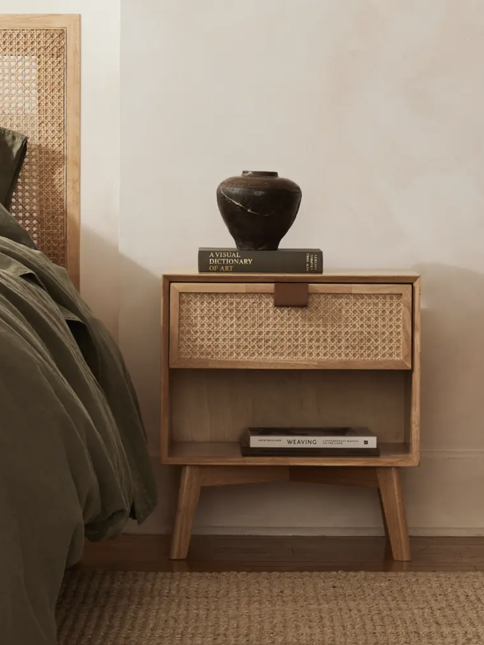 A wooden bedside table with a woven front, two books, and a dark ceramic vase is beside a bed with green bedding. A book titled "Weaving" is visible on the lower shelf.