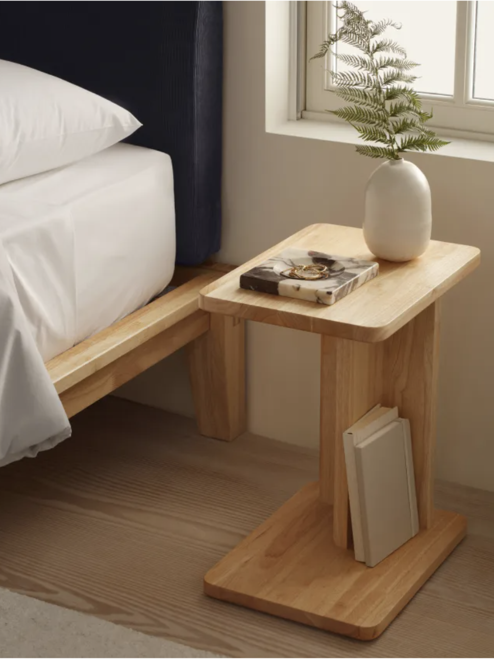 A wooden bedside table with a book and glasses on top, a vase with a fern, and a book stored in its base, next to a bed.