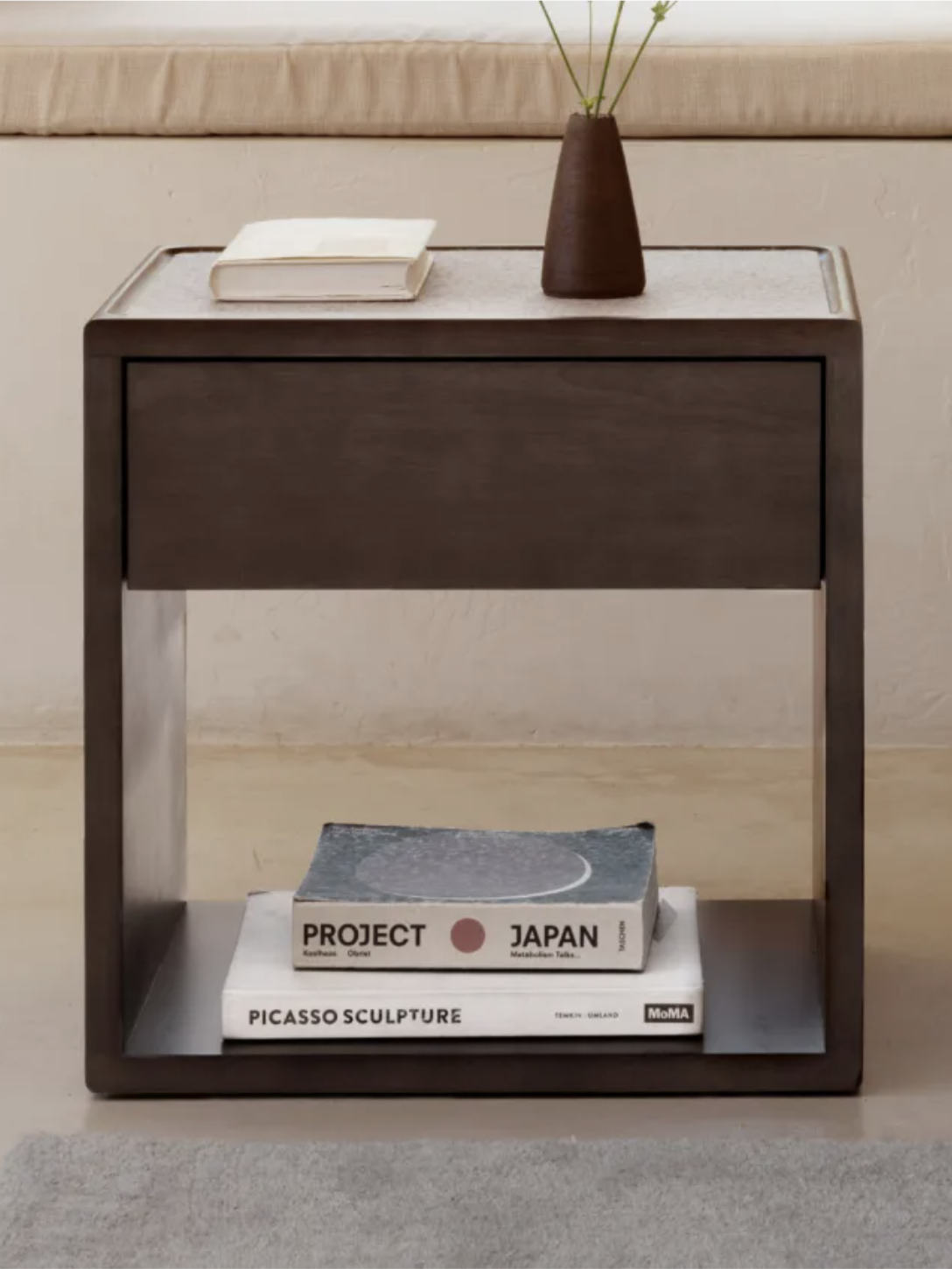 A small wooden side table with a drawer, holding a vase and books. Books include "Project Japan" and "Picasso Sculpture." A cushion sits on the window ledge above the table.
