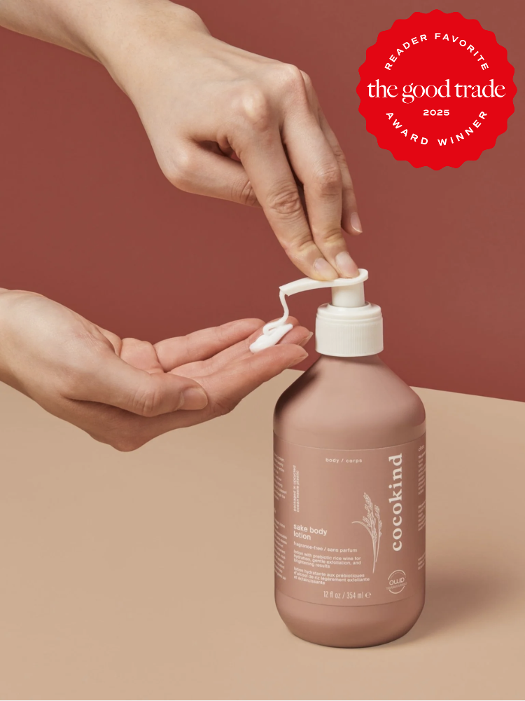 A hand presses the pump of a Cocokind bottle, dispensing lotion onto the other hand. The background is a two-tone of muted red and beige. A red award seal is in the top right corner.