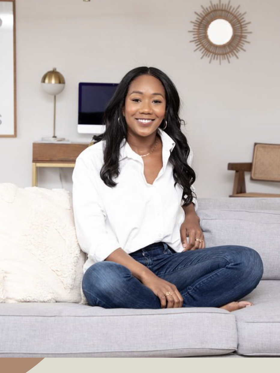 A woman in a white shirt and jeans sits cross-legged on a light gray sofa, smiling. A desk with a computer and lamp are in the background.