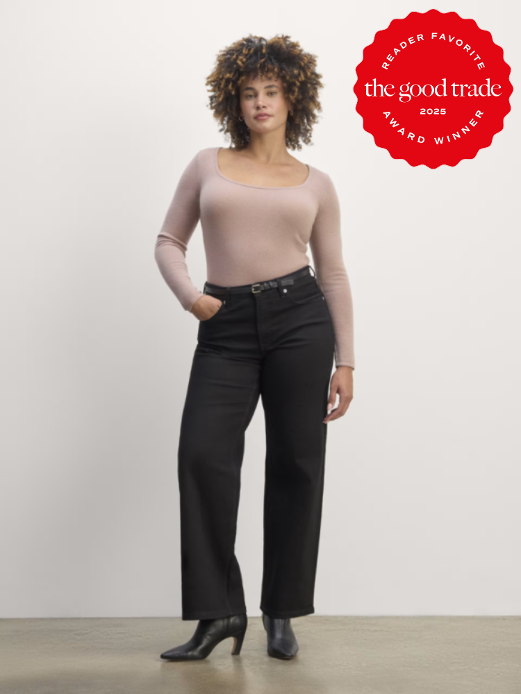 Person standing in front of a white background wearing a light pink top, black pants, and black boots. A red award badge is on the top right corner.