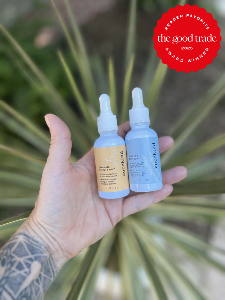 A tattooed hand holding two bottles of eco-oil over blurred greenery. One bottle is labeled "Calm Down" and the other "Glow Brighter." A red badge reads "Reader Favorite, The Good Trade, 2025 Award Winner.