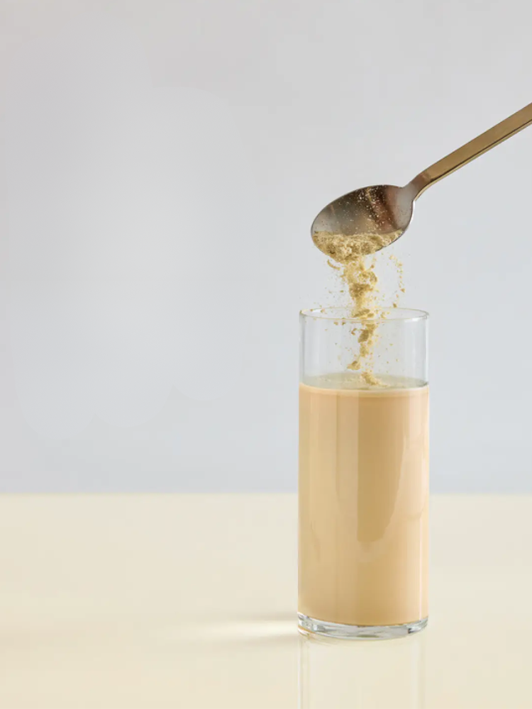 A spoonful of powder is being added to a glass of light brown liquid on a beige surface with a plain background.