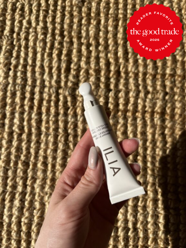 A hand holding a tube of ILIA product against a woven surface. Red badge in the corner reads: "Reader Favorite, The Good Trade, 2025 Award Winner.