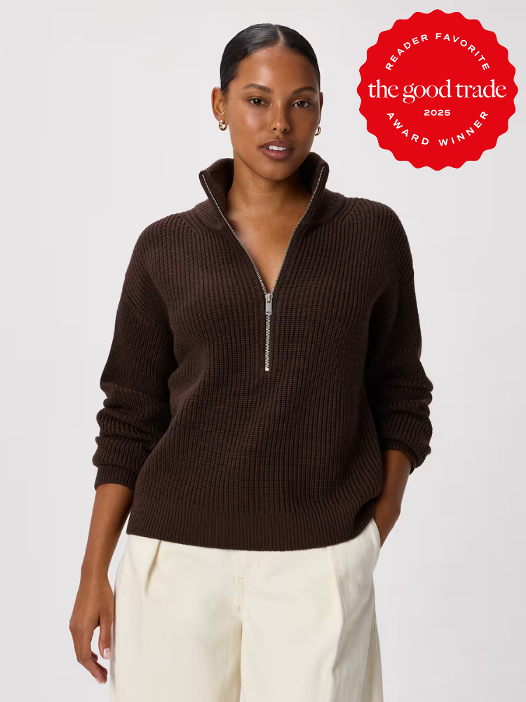 A woman wears a dark brown ribbed half-zip sweater and cream pants, standing against a plain background. A red "The Good Trade Award Winner 2025" badge is in the top right corner.