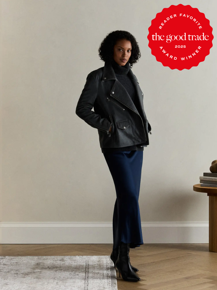 A woman stands indoors wearing a black jacket, dark turtleneck, blue satin skirt, and black boots. A "The Good Trade 2025 Award Winner" badge appears in the top right corner.