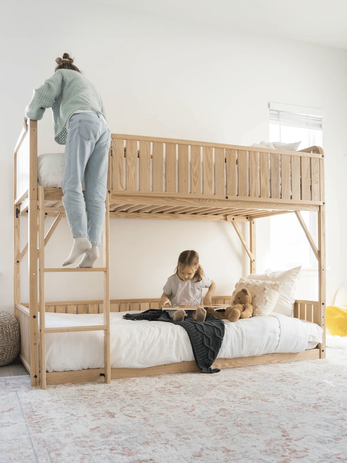 Two children use a wooden bunk bed; one climbs the ladder to the top bunk while the other sits on the lower bunk, reading a book next to a teddy bear.
