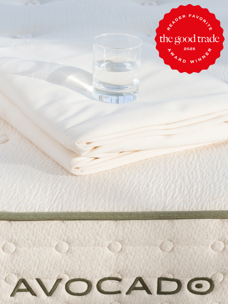 A glass of water on folded white linens atop an Avocado mattress. A red badge reads "Reader Favorite, The Good Trade Award Winner 2025.
