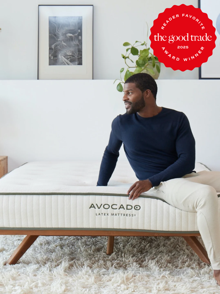 A man sits on an organic Avocado Latex Mattress in a minimalist room adorned with a shag rug, artwork, and lush greenery. The image proudly displays a "Reader Favorite" award badge.