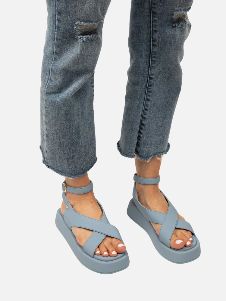 Person wearing distressed blue jeans and light blue platform sandals with crisscross straps, standing on a white background.