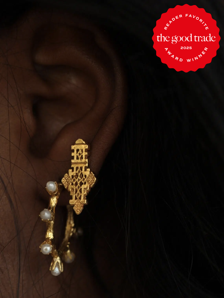 Close-up of a person's ear wearing ornate gold earrings with pearl details. The top right corner shows a red badge with "Reader Favorite" and "The Good Trade 2025 Award Winner" text.
