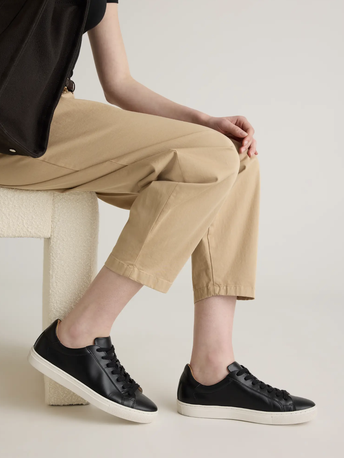Person sitting on a beige chair wearing black sneakers, tan pants, and a black shirt, holding a bag.