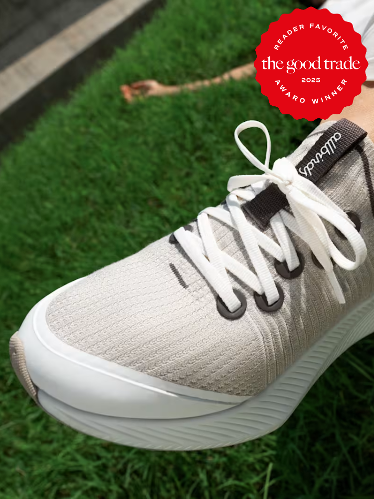 Close-up of a person wearing light gray sustainable sneakers on grass, with a red "The Good Trade 2025 Reader Favorite Award Winner" badge in the upper right corner.