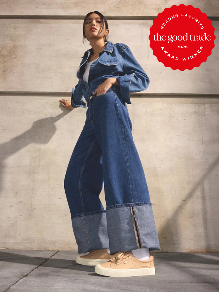 Woman wearing a denim jacket, wide-leg jeans, and sustainable sneakers stands against a concrete wall; "The Good Trade 2025 Award Winner" badge appears in the top right corner.