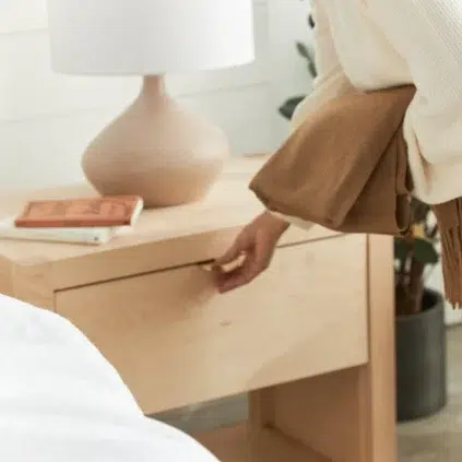 Person holding a brown sweater opens a wooden nightstand drawer beside a table lamp and book.