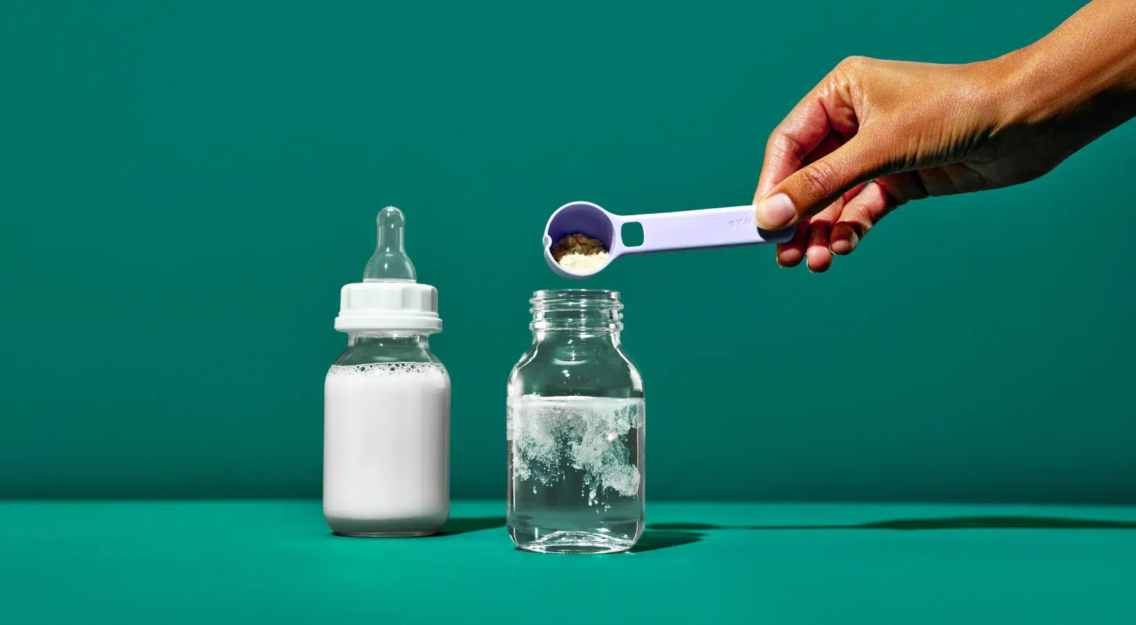 A hand pours powdered formula into a jar of water beside a filled baby bottle, all set against a solid teal background.