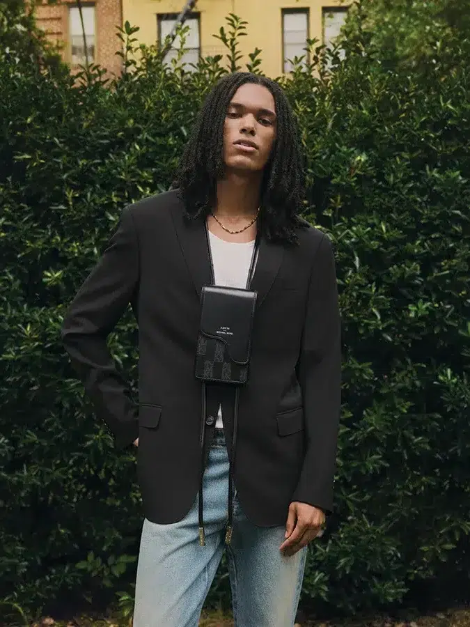 Person with long hair wearing a black blazer, white shirt, and jeans, stands in front of dense green foliage, with a crossbody bag hanging in front.