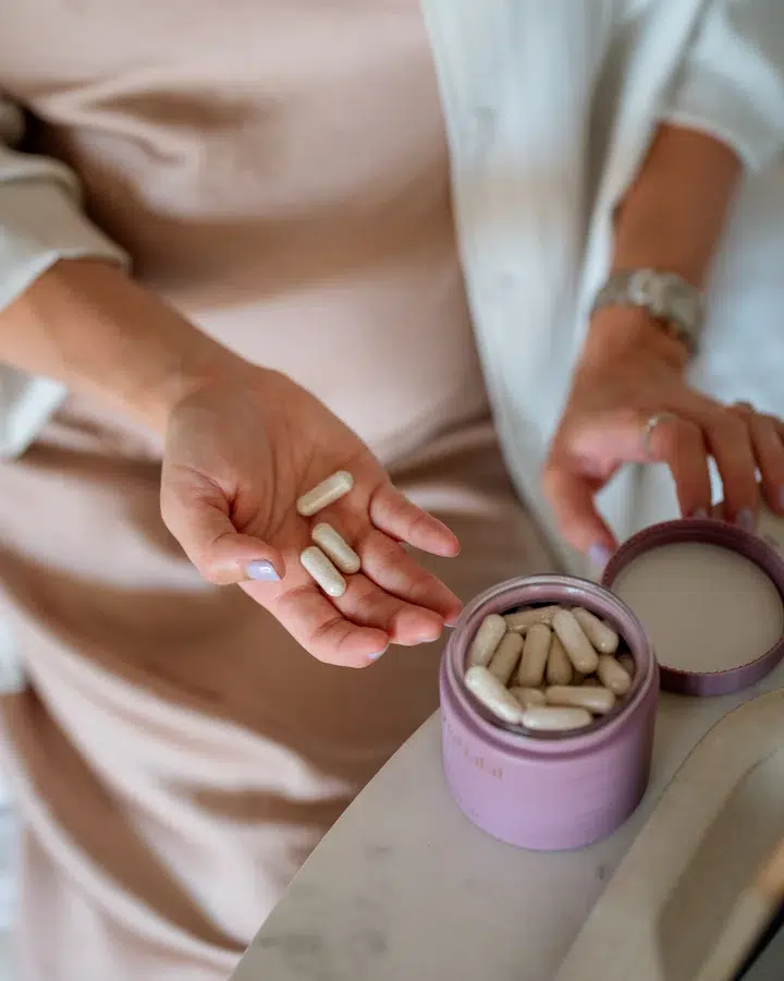 A person holds several white prenatal vitamin capsules in one hand while standing next to an open container filled with more capsules on a table.