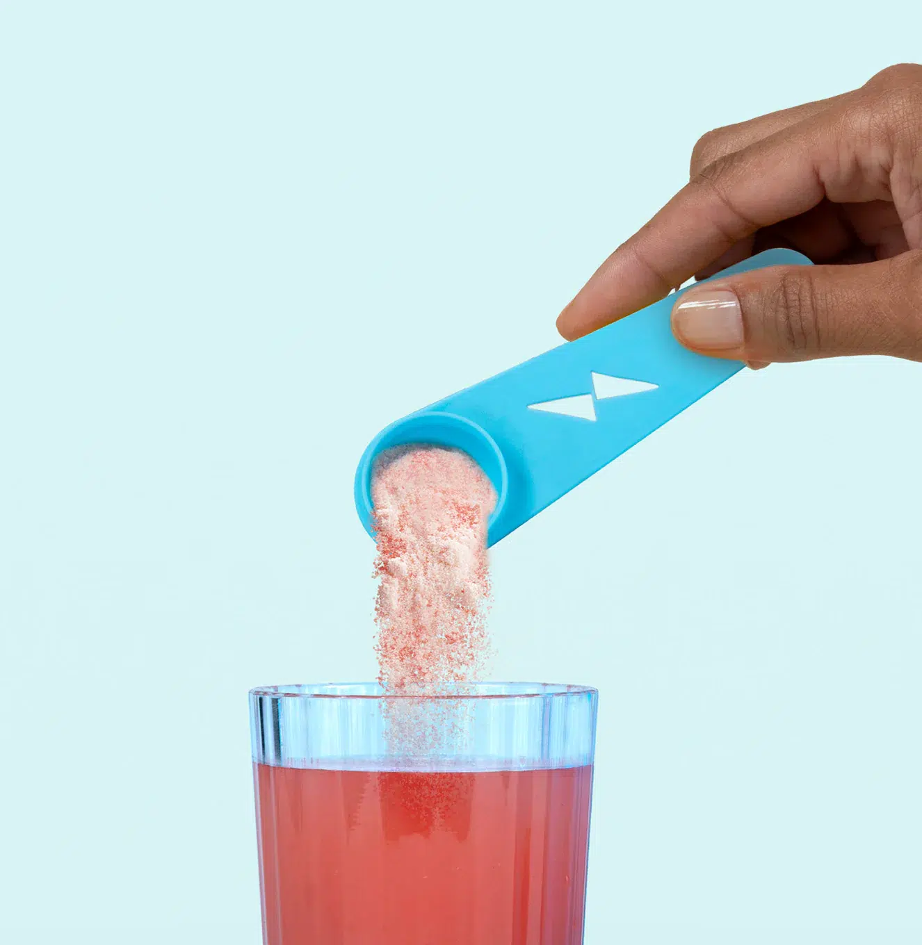 A hand pours pink powder from a blue scoop into a glass filled with red liquid against a light blue background.