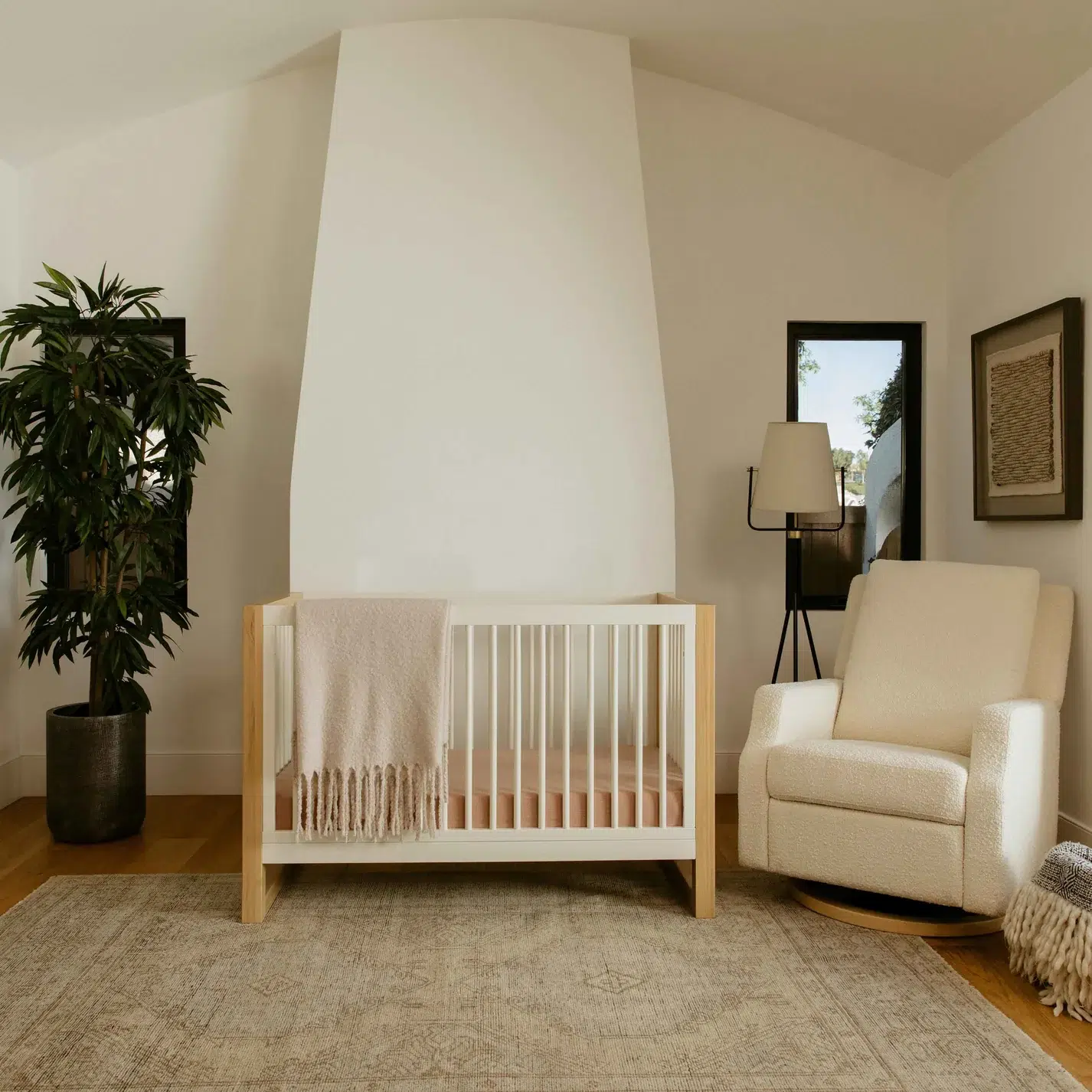 A modern nursery features a white crib with a beige blanket, a cream armchair, a tall potted plant, a rug, and minimal wall decor in a softly lit room.